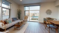 This single-floor residence showcases a thoughtfully designed contemporary interior with a cohesive neutral palette and warm wood-look flooring throughout. The open-concept kitchen is a standout feature, equipped with sleek two-tone cabinetry in dark charcoal and muted blue-grey, a white subway tile backsplash, light quartz countertops, and a full suite of stainless steel appliances including a range, over-the-range microwave, dishwasher, and French door refrigerator. Under-cabinet lighting adds a polished finishing touch. Adjacent to the kitchen, the dining area benefits from tall ceilings and large floor-to-ceiling windows that draw in generous natural light and offer views of the surrounding neighborhood. The living room continues this bright, airy quality with an expansive sliding glass window wall overlooking city rooftops and hills. The bedroom is well-proportioned, featuring wide windows with elevated views and clean white walls. Two bathrooms mirror each other in design, each featuring a tub-shower combination with sliding glass enclosure, light tile flooring, a white quartz vanity, and warm wood cabinetry, creating a calm and functional retreat.
