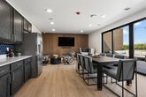 LOT Skydeck is a 500-square-foot rooftop amenity space thoughtfully designed to blend indoor comfort with open-air enjoyment. The interior is divided into a lounge area, a fully equipped kitchen, and a compact dining nook. The lounge features a striking wood slat accent wall paired with a wall-mounted TV, complemented by contemporary patterned sofas, a round black coffee table, and coastal-inspired wall art, creating a refined yet relaxed atmosphere. The kitchen is outfitted with rich dark wood cabinetry, a bold blue hexagonal tile backsplash, white countertops, a stainless steel refrigerator, and light wood-style flooring — offering full functionality for entertaining and catering. The dining nook provides an intimate seating area with a round pedestal table, modern chairs, and decorative artwork. A set of floor-to-ceiling glass doors opens directly onto the expansive outdoor deck, which is finished with large-format tile pavers and lined with multiple dining tables and chairs arranged for group gatherings. Overhead string lights extend across the deck, while a black metal railing frames sweeping open views of the surrounding landscape under open skies, making this rooftop an ideal venue for events and social gatherings.