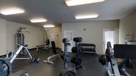 This fitness center is a dedicated single-floor space designed to support a wide range of strength training and exercise activities. The room features a clean, neutral color palette with light gray walls and a dark rubberized flooring that provides both durability and safety during workouts. Bright overhead fluorescent lighting illuminates the entire space evenly, ensuring excellent visibility throughout.

The facility is well-stocked with a variety of free weights, including a fully organized dumbbell rack holding an extensive range of weights, as well as multiple barbells and weight plates positioned on upright storage racks. Several adjustable workout benches are arranged throughout the room, accommodating exercises from flat to incline positions. A cable resistance machine is situated toward the back of the space, offering additional exercise variety.

Instructional fitness charts are mounted on the walls, providing users with exercise guidance and reference material. The space also includes a designated office area with a frosted glass door, suggesting on-site management or staff support. A secondary door on the far wall provides additional access to the space. The overall layout is open and functional, allowing for easy movement between equipment stations and accommodating multiple users simultaneously.