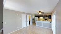 This two-floor residence offers a practical and well-appointed living environment with a clean, functional layout. The main living area features light-toned ceramic tile flooring that extends seamlessly throughout, creating a cohesive and easy-to-maintain surface underfoot. A ceiling fan with integrated lighting anchors the open living space, providing both airflow and illumination.

The kitchen is positioned in an open-concept arrangement, separated from the main living area by a raised breakfast bar or peninsula finished in white. Dark espresso-toned cabinetry lines the upper and lower kitchen walls, providing ample storage and a contrasting design element. The kitchen is equipped with a white refrigerator, electric range, and under-cabinet range hood, complemented by track lighting overhead for focused task illumination.

Adjacent to the main living space, two interior doors lead to additional rooms — one appears to open into a utility or laundry area, visible by the appliances within. The neutral gray wall tones throughout give the space a modern, understated feel that complements a wide range of furnishing styles. The overall layout is open and airy, making efficient use of the available space across both floors.