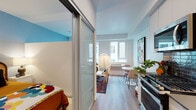 This well-designed single-floor unit showcases a thoughtful, open layout that maximizes functionality within a compact footprint. Light wood-tone flooring runs continuously throughout, creating a cohesive and airy feel across all areas.

The sleeping area is separated from the main living space by a frosted sliding glass partition, offering privacy while maintaining an open visual connection. A vibrant blue accent wall adds a bold design element to this zone.

The living area features large windows that allow generous natural light to fill the space, with views of the surrounding urban streetscape. A wall-unit HVAC system provides climate control, and the room comfortably accommodates a seating area and small dining table.

The kitchen is outfitted with white flat-front cabinetry, light stone countertops, a stainless steel undermount sink, a gas range, built-in microwave, and a sleek dark tile backsplash that adds contrast and texture.

The bathroom is clean and modern, featuring a white vanity with a dark countertop, an undermount sink, a large mirror, and a toilet. The space is well-lit and finished with the same light wood flooring found throughout the unit.