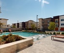 This contemporary residential complex in Seagoville, TX, showcases a harmonious blend of modern architecture and inviting amenities. The main building boasts a striking facade of light-colored stone with large windows, including triangular ones that add architectural interest. A metal roof complements the stone exterior, creating a sleek, upscale appearance.

The property's centerpiece is an expansive pool area, featuring a large, crystal-clear swimming pool surrounded by a spacious deck. Adjacent to the pool, a covered outdoor living area provides shade and comfort, complete with seating and dining furniture. This space is perfect for relaxation and socializing.

The complex comprises multiple residential buildings, varying in height and design, creating a diverse and dynamic community atmosphere. Landscaping elements, including trees and well-maintained shrubbery, add natural beauty to the surroundings.

Safety features include a perimeter fence and ample lighting throughout the common areas. The complex also offers parking facilities for residents and guests. With its modern amenities and thoughtful design, this Seagoville property provides a resort-like living experience, combining comfort, style, and community in a suburban Texas setting.