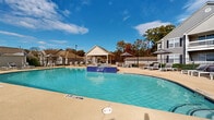 Located at 6229 Thomaston Road in Macon, Georgia, this outdoor pool and recreation area offers a well-appointed amenity space designed for relaxation and leisure. The centerpiece is a generously sized swimming pool with crystal-clear blue water, framed by a smooth concrete sundeck that provides ample room for lounging. A built-in spa with blue tile accents sits prominently within the pool area, serving as a distinctive architectural focal point.

The deck is furnished with numerous chaise lounge chairs and patio tables shaded by market umbrellas, offering comfortable spots to relax throughout the day. A charming covered pavilion with a pitched roof provides a shaded gathering area, complemented by additional outdoor seating nearby. A dedicated pool house building with an arched window and navy blue entry door serves as a functional support structure for the amenity area.

The space is enclosed by a wrought iron fence, maintaining both security and an open, airy feel. Mature trees with seasonal foliage line the perimeter, providing a natural backdrop and additional shade. Decorative lamp posts are positioned throughout the space, ensuring the area is well-lit during evening hours. The overall ambiance is reminiscent of a resort-style retreat, offering a refined and inviting outdoor experience.