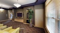This single-floor screening room is thoughtfully designed to deliver a premium home theater experience in a communal setting. The main viewing area is furnished with rows of generously sized, cream-toned leather armchairs arranged to face a large flat-screen display mounted within a custom built-in media console along the front wall. Each seat is paired with a sleek, circular chrome side table, adding a polished, contemporary touch.

The walls are finished in warm taupe tones and adorned with tall, vertical acoustic panels illuminated by elegant wall sconces, enhancing both the room's sound quality and visual sophistication. A deep purple crown molding runs along the ceiling perimeter, providing a rich contrast to the lighter walls and contributing to the room's cinematic atmosphere. Recessed ceiling lights offer adjustable ambient illumination throughout.

At the rear of the space, a comfortable seating alcove features additional leather chairs and a gallery-style arrangement of framed artwork, creating a welcoming transition zone. A built-in niche showcases a tall indoor plant, adding a natural, organic element to the décor. Natural light filters in through slatted window blinds along one wall, while patterned carpet in deep earth tones grounds the entire space and reinforces the room's upscale, theater-inspired aesthetic.