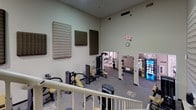 This well-appointed, two-level fitness center offers a thorough workout environment designed to accommodate a wide range of exercise needs. The main floor is expansive, featuring soaring double-height ceilings that create an open, airy atmosphere. Large acoustic panels are mounted along the walls, contributing to a more controlled sound environment during workouts. The ground level is generously outfitted with a variety of strength training equipment, including multiple Paramount and Matrix cable and resistance machines, adjustable benches, and a dumbbell rack, all arranged across durable, carpet-covered flooring with rubberized zones. A staircase leads to the mezzanine level above, which houses additional cardio equipment including Matrix treadmills and stationary bikes, set on protective rubber flooring and illuminated by recessed skylight panels. The upper level is open to the floor below, reinforcing the sense of vertical space throughout the facility. Adjacent to the main workout area, a vending area with snack and beverage machines provides convenient refreshments. The facility is well-lit throughout with a combination of overhead fluorescent and recessed lighting, and security cameras are present for added safety. The overall ambiance is functional, clean, and purpose-built for fitness.