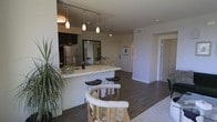 This single-floor space showcases a thoughtfully designed open-concept layout that seamlessly connects the kitchen, dining, and living areas into one cohesive and inviting environment. The kitchen is outfitted with dark cabinetry, stainless steel appliances, and a sleek white breakfast bar that doubles as a casual dining space, complete with modern bar stools. A set of elegant pendant lights hang above the bar, complementing the track lighting overhead and adding a warm, layered ambiance to the space. Rich, dark hardwood-style flooring runs throughout, grounding the interior with a sense of warmth and continuity. The living area is open and airy, benefiting from the fluid connection to the kitchen and dining space. Neutral white walls keep the atmosphere bright and versatile, while a ceiling fan in the living area adds both function and a subtle decorative touch. A large mirror in the kitchen area reflects light and visually expands the space. Hallways lead to additional private rooms, hinting at a well-organized floor plan. The overall aesthetic is clean, contemporary, and functional, making excellent use of the available space with intentional design choices throughout.
