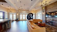 Located at 602 Laconia Cir in Spartanburg, SC, this community clubhouse at Auder Living offers a thoughtfully designed single-floor layout that combines modern aesthetics with industrial-chic character. The main living area greets visitors with soaring ceilings, an eye-catching large sculptural art installation, and clusters of Edison bulb pendant lights that cast a warm, inviting glow. Plush tan leather sofas anchor the lounge space, while an exposed brick accent wall adds rich texture and depth. A second seating area features sleek mid-century modern leather chairs arranged around a wooden coffee table, with a wall-mounted TV set against the same signature brick backdrop. The combined dining, kitchen, and living area is a standout feature, boasting a large white quartz island with industrial metal barstools, exposed wood ceiling beams, corrugated metal accents, open shelving, and a dedicated coffee station. The hallway area provides a quiet retreat with swivel lounge chairs and a wall-mounted TV. The game room is a vibrant social hub, highlighted by a bold purple accent wall, a foosball table, a billiard table, and abundant natural light from large windows. Throughout, polished concrete floors, eclectic artwork, and carefully curated lighting fixtures unify the space with a contemporary, community-forward atmosphere.