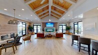Located at 4625 Gordon Rd in Wilmington, NC, this single-story community clubhouse showcases a thoughtfully designed coastal aesthetic throughout its well-appointed spaces. The centerpiece of the facility is a grand lounge area distinguished by dramatic vaulted ceilings with warm wood tongue-and-groove paneling framed by white structural beams, recessed lighting, and ceiling fans. This expansive gathering space features light-toned hardwood flooring, wood-clad accent walls, a sleek linear fireplace topped by a mounted flat-screen TV, and generous seating arrangements. Multiple sets of black-framed glass doors flood the interior with natural light and provide views of the surrounding outdoor areas. An adjacent lounge nook offers additional casual seating beneath stylish pendant lighting, coastal-inspired artwork, and a wood-paneled feature wall. The coworking area provides a large conference table, individual booth-style workstations, and a service counter with a printer and beverage station. The full kitchen is equipped with stainless steel appliances, double wall ovens, a large center island with seating, and a vibrant turquoise tile backsplash. Additional amenities include restrooms and a water station accented by a decorative gold mosaic tile wall.