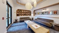 Located at 2530 NE 181st St in North Miami Beach, FL, this stylish library lounge offers a refined and inviting atmosphere designed for comfort and leisure. The single-room space is characterized by soaring ceilings adorned with a trio of oversized paper lantern pendant lights that cast a warm, ambient glow throughout the room. A distinctive tray ceiling with warm wood trim frames the space elegantly, adding architectural depth and visual interest.

The room features a built-in open shelving unit with arched detailing, displaying a curated selection of decorative objects, ceramics, and books. Opposite the shelving, a recessed wall niche showcases a gallery-style arrangement of framed New Yorker magazine covers, illuminated by a sleek brass picture light, lending a cultural and artistic touch to the space.

Seating is plentiful and thoughtfully arranged, with multiple plush velvet sofas, tufted lounge chairs, and upholstered benches positioned around low wooden coffee tables atop a patterned blue-grey carpet. Brass wall sconces provide additional accent lighting. A set of black-framed glass double doors opens to an outdoor balcony, inviting natural light and views of the surrounding property. The overall ambiance is warm, sophisticated, and well-suited for quiet reading or social gathering.