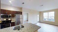 This single-floor residence offers a thoughtfully designed open-concept layout that seamlessly connects the kitchen and living areas. The kitchen is a standout feature, showcasing granite countertops with a built-in sink on a peninsula-style breakfast bar that extends into the main living space, providing both functionality and a natural gathering point. Rich dark-stained cabinetry contrasts elegantly with the light granite surfaces, while a full-size stainless steel refrigerator and coordinating appliances complete the modern aesthetic. Light hardwood flooring runs throughout the space, adding warmth and visual continuity from room to room. The living area benefits from natural light streaming through windows and a glass-panel door, creating a bright and welcoming atmosphere. Neutral warm-toned walls provide a clean backdrop that complements the overall palette. A doorway leads to an additional room, offering flexible use as a bedroom or secondary living space. The space features recessed and track lighting, and ceiling vents indicate a central HVAC system. With its clean lines, quality finishes, and open flow, this residence delivers a practical yet polished living environment.