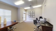 This single-floor professional office space offers a polished and functional environment suited for business operations. The space is finished with neutral taupe walls, crown molding, and textured wall-to-wall carpeting throughout, lending a clean, cohesive aesthetic. Recessed fluorescent lighting ensures consistent, bright illumination across all areas.

The main office area features a substantial dark wood executive desk accompanied by ergonomic rolling chairs, providing ample workspace. Multiple windows fitted with white horizontal blinds allow natural light to filter in, contributing to an open and comfortable atmosphere. A framed nine-panel gallery wall displaying black-and-white architectural photography serves as a distinctive decorative focal point.

A dedicated business center alcove along one wall houses a dark granite-topped workstation equipped with two desktop computers, keyboards, and a shared laser printer — ideal for document management and general computing needs. A glass-paneled interior door with transom adds architectural character while maintaining visual connectivity between areas.

The space also includes an additional seating area with rolling chairs positioned near the workstations, offering flexibility for collaborative use. Security is supported by a wall-mounted camera. The overall design balances functionality with a refined, professional aesthetic, making this space well-suited for leasing office or business center use.