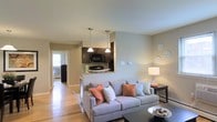 Located at 239 W Johnson Street in Philadelphia's Germantown neighborhood, this single-floor unit offers a thoughtfully designed open-concept layout that connects the living and kitchen areas into one cohesive space. The living area features warm hardwood flooring, neutral-toned walls, and ample room for comfortable furnishings. A raised peninsula with granite countertop serves as the natural dividing point between the living space and kitchen, providing both a functional workspace and a casual seating area. The kitchen is outfitted with dark espresso cabinetry, black appliances including a built-in microwave, and two pendant lights that hang above the peninsula, adding a stylish focal point to the space. The open sightlines from the living area through to a hallway, where natural light filters in from a window beyond, contribute to an airy, well-lit atmosphere throughout. The consistent hardwood flooring flows throughout the unit, reinforcing a sense of continuity. The neutral color palette and clean architectural lines create a versatile, move-in-ready environment well-suited for a range of interior styles.