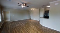 This single-floor space offers a thoughtfully designed open-concept layout that combines the living and kitchen areas into one cohesive, flowing environment. Rich hardwood-style flooring extends throughout, lending warmth and continuity to the space. The living area is highlighted by elegant crown molding, a ceiling fan with light fixture for year-round comfort, and recessed track lighting that provides both ambiance and functionality. Neutral, soft-toned walls create a clean and versatile backdrop adaptable to a variety of interior styles. The kitchen is partially open to the living area, separated by a curved breakfast bar topped with a granite-style countertop that adds a refined touch. A black refrigerator and additional built-in appliances are visible, complementing the kitchen's practical and modern aesthetic. White raised-panel cabinetry adds classic character to the culinary space. A doorway visible from the main living area leads to additional rooms, suggesting a well-organized floor plan. The overall ambiance is bright, open, and move-in ready, with tasteful architectural details that elevate the everyday living experience throughout this single-level space.