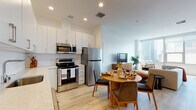 Located at 3348 Wilshire Blvd in the heart of Los Angeles, this contemporary single-floor residence offers a thoughtfully designed open-concept layout that seamlessly connects the kitchen, dining, and living areas. The kitchen is outfitted with full stainless steel appliances — including a refrigerator, electric range, over-the-range microwave, and dishwasher — complemented by crisp white cabinetry, white quartz countertops, a tile backsplash, and light hardwood-style flooring that flows throughout the unit. The dining area accommodates a round table with seating, positioned between the kitchen and living space for easy entertaining. The living area is bright and airy, benefiting from large floor-to-ceiling windows that frame sweeping views of the Los Angeles skyline, including notable landmarks. The bedroom is generously proportioned, also featuring expansive windows with city views, and connects to a well-appointed bathroom complete with a single-sink vanity with stone countertop, a soaking tub with shower, and travertine-style tile surround. Clean white walls, recessed lighting, and consistent flooring throughout lend the space a cohesive, modern aesthetic ideal for urban living in the Koreatown/Midtown LA corridor.