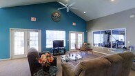 This single-floor space makes a striking first impression with its dramatic vaulted ceilings that rise to a sharp peak, creating an expansive, airy atmosphere. A bold teal accent wall serves as the room's focal point, adorned with a decorative wooden tree medallion that adds a natural, artistic touch. A ceiling fan is mounted at the apex, ensuring comfortable airflow throughout the space.

The living area is generously appointed with plush, cushioned seating arranged around a glass-top coffee table, making it well-suited for relaxed gatherings. Natural light pours in abundantly through multiple large windows and a set of French doors, which open to an outdoor area, seamlessly blending indoor and outdoor living.

A dedicated media area features a flat-screen television on a classic entertainment stand, while a separate dining nook with wrought-iron chairs and a small table provides an intimate spot for casual meals or conversation. The neutral carpeted flooring complements the warm, welcoming color palette of the walls.

Overall, the space strikes a balance between comfort and style, with its high ceilings, vibrant color choices, and plentiful natural light contributing to an inviting and functional environment suited for community use or social gatherings.