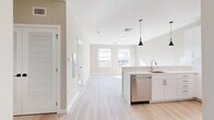 This thoughtfully designed 1-bedroom, 1-bathroom unit offers a clean, modern aesthetic throughout its single-floor layout. The open-concept kitchen and living area serves as the heart of the home, featuring crisp white cabinetry, a large quartz waterfall island, stainless steel appliances including a refrigerator, range, microwave, and dishwasher, and a sleek single-basin sink — all illuminated by stylish black pendant lighting. Light hardwood flooring flows seamlessly throughout, enhancing the bright and airy feel of the space. The living area benefits from multiple windows that invite an abundance of natural light and offer views of the surrounding neighborhood. The spacious bedroom is well-appointed with a generous closet, light-toned walls, and large windows that frame outdoor views and fill the room with sunlight. A second, additional room provides flexibility for use as a home office or sitting area, also featuring a bright window. The bathroom is finished with a clean, modern palette, including a white vanity with a quartz countertop, gold-toned hardware, a framed mirror, and marble-style tile flooring. A louvered closet door near the entry adds a functional storage element to this polished, move-in-ready unit.