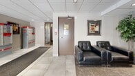 The lobby at 450 Victoria Ave serves as the primary entrance and welcoming space for the building, offering a clean, functional, and well-maintained environment for residents and visitors alike. Large floor-to-ceiling glass doors and sidelights at the main entrance flood the space with natural light while providing clear views of Victoria Avenue and the surrounding streetscape. The flooring features large-format white ceramic tiles, complemented by a durable grey carpet runner leading from the entrance, creating a practical and polished aesthetic. A drop ceiling with recessed lighting and a white tile grid provides consistent, bright illumination throughout. One wall is dedicated to an extensive bank of individual resident mailboxes, offering convenient mail access directly from the lobby. Canada Post parcel lockers are also present, providing secure package delivery solutions. A bulletin board displays building notices and community information. A comfortable seating area, furnished with black leather sofas, a wooden coffee table, and decorative elements including framed artwork and an indoor plant, creates a relaxed waiting space. Baseboard heating units along the perimeter walls ensure year-round comfort. A stairwell door provides access to the rest of the building, rounding out this practical and welcoming lobby.