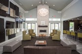 Located in Franklin, Tennessee, this single-floor community clubhouse offers a thoughtfully designed gathering space that blends contemporary style with comfortable functionality. The main lounge area makes an immediate impression with its soaring double-height ceilings, which amplify the sense of openness and allow natural light to filter in through tall glass entry doors. Light wood-look tile flooring runs throughout, complemented by area rugs that define distinct seating zones.

The lounge features multiple conversation areas furnished with patterned wingback chairs in bold gold tones, plush sofas, and upholstered ottomans arranged around a wooden coffee table. A dramatic accent wall serves as the room's focal point, displaying an artistic grid of dark espresso and mirrored panels surrounding a wall-mounted flat-screen TV — an eye-catching design element that adds texture and depth. A stone fireplace anchors another corner of the space, adding warmth and architectural character.

Adjacent to the lounge is a well-appointed kitchenette area, finished with dark espresso cabinetry, granite-style countertops, open shelving, and a striking metallic hexagonal tile backsplash. A breakfast bar with seating and a mounted TV adds to the area's utility. Recessed lighting throughout ensures the space is well-lit and inviting, making this clubhouse an ideal setting for both relaxation and social gatherings.