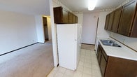 This single-floor space offers a straightforward and functional layout, with the kitchen serving as a central feature. The kitchen is equipped with dark brown cabinetry mounted above the refrigerator and along the walls, providing ample storage. Light-toned tile flooring runs throughout the kitchen area, offering easy maintenance and a clean aesthetic. A full-size white refrigerator anchors the kitchen, accompanied by a washer nearby, a sink integrated into dark cabinetry, and an adjoining countertop workspace. A decorative wallpaper border runs along the upper walls, adding a subtle touch of character to the space. The kitchen transitions seamlessly into the adjacent carpeted living area, which features neutral-toned carpet and white walls, creating a bright and open feel. A hallway visible from the kitchen leads to additional rooms, including a space with a closet featuring built-in shelving, suggesting a bedroom or storage area. The textured ceiling is consistent throughout, and recessed lighting in the kitchen ensures adequate illumination. Overall, the space presents a practical, no-frills interior suited for everyday living.