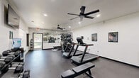 Located at 2450 Centennial Ridge Way in Raleigh, NC, this fitness center offers a well-appointed, single-floor exercise space designed to support a comprehensive workout routine. The room features durable dark rubber flooring throughout, white walls, and recessed lighting supplemented by dual black ceiling fans that ensure consistent airflow and comfort during workouts. A large wall mirror on one end visually expands the space and aids in form checking during exercise. Cardio equipment includes multiple treadmills, a stair climber, and a stationary bike with an integrated display screen. Strength training options are equally thorough, with a multi-rack dumbbell set spanning a wide weight range, kettlebells, medicine balls, a flat weight bench, a cable machine, and TRX suspension straps. A wall-mounted flat-screen television provides entertainment, while a water cooler and waste bin add everyday convenience. Natural light filters in through several windows fitted with horizontal blinds, and a glass-panel entry door offers a view to the outdoors. Motivational fitness posters and a chalk-style message board contribute to an energizing atmosphere. The space is clean, modern, and efficiently organized to maximize usability.