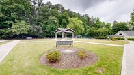 This expansive outdoor community space offers a thoughtfully designed landscape that balances open green areas with intimate gathering spots. Wide, smooth concrete walkways wind throughout the grounds, connecting the various amenities and encouraging leisurely strolls. A central covered pavilion with a pitched roof and sturdy timber construction serves as a natural focal point, providing a shaded retreat within the open lawn. Nearby, a dedicated fire pit area features a circular arrangement of Adirondack chairs set into a mulched landscape bed, creating an inviting spot for relaxed gatherings. A bocce ball court adds a recreational element to the grounds, complemented by well-placed park benches that offer additional seating throughout the space. Manicured shrubs, ornamental trees including crape myrtles, and lush turf lawns contribute to the polished aesthetic, while a dense backdrop of mature trees provides natural privacy and a verdant sense of enclosure. A gated entry with a small gatehouse structure marks a formal entrance to the property, reinforcing the well-kept and secure character of the grounds. Additional amenities visible in the distance include a pool area, further enhancing the range of outdoor recreational offerings available across these community grounds.