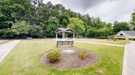 This expansive outdoor community space offers a thoughtfully designed landscape that balances open green areas with intimate gathering spots. Wide, smooth concrete walkways wind throughout the grounds, connecting the various amenities and encouraging leisurely strolls. A central covered pavilion with a pitched roof and sturdy timber construction serves as a natural focal point, providing a shaded retreat within the open lawn. Nearby, a dedicated fire pit area features a circular arrangement of Adirondack chairs set into a mulched landscape bed, creating an inviting spot for relaxed gatherings. A bocce ball court adds a recreational element to the grounds, complemented by well-placed park benches that offer additional seating throughout the space. Manicured shrubs, ornamental trees including crape myrtles, and lush turf lawns contribute to the polished aesthetic, while a dense backdrop of mature trees provides natural privacy and a verdant sense of enclosure. A gated entry with a small gatehouse structure marks a formal entrance to the property, reinforcing the well-kept and secure character of the grounds. Additional amenities visible in the distance include a pool area, further enhancing the range of outdoor recreational offerings available across these community grounds.