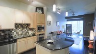 This single-floor space showcases an open-concept layout that seamlessly connects the kitchen and living areas, creating a cohesive and functional environment. The kitchen is a standout feature, highlighted by sleek dark granite countertops on a curved island that serves as both a workspace and casual gathering spot. Light maple cabinetry provides ample storage, while a classic checkered tile backsplash adds visual interest behind the sink. Stainless steel appliances, including a built-in wall oven, microwave, and a smooth cooktop integrated into the island, give the kitchen a polished, contemporary feel. Track lighting and pendant fixtures suspended from the notably high ceilings ensure well-distributed illumination throughout. The living area, visible beyond the kitchen island, is bathed in natural light streaming through large windows. Ceiling fans in both zones contribute to the overall comfort of the space. The open floor plan fosters a spacious ambiance, making the transition between cooking, dining, and relaxing effortless. The combination of modern finishes, elevated ceilings, and abundant natural light gives this space a bright, urban character.
