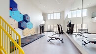 This two-story fitness studio in Louisville, KY offers a well-rounded workout environment spread across two distinct levels, each designed to accommodate a variety of exercise disciplines. The main floor is the architectural centerpiece of the space, boasting soaring double-height ceilings that flood the area with natural light through large clerestory windows positioned near the top of the walls. A bold hexagonal climbing wall in blue, tan, and cream tones anchors one side of the room, paired with protective floor mats below. Full-length mirrors line an adjacent wall, visually expanding the space while supporting strength training and form work. The main floor also houses a multi-use cable machine, adjustable benches, a dumbbell rack, and a functional training area. A door on this level provides direct access to an outdoor area. A yellow-railed staircase leads to the upper level, where light wood-look flooring sets a warmer tone. This floor features treadmills and ellipticals positioned beside large windows overlooking the surrounding neighborhood, as well as recumbent bikes, upright cycles, and a rowing machine. A wall-mounted television and water station complete the upper level's amenities, creating a bright, practical space for low-impact cardio training.