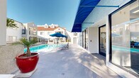 Located at 333 Andover Dr in Burbank, California, this outdoor amenity space is designed to maximize the region's abundant sunshine and mild climate. The centerpiece is a large, crystal-clear swimming pool surrounded by a generously sized concrete sundeck furnished with multiple blue lounge chairs, perfect for relaxing under the Southern California sun. A separate spa with tiled surround provides an additional area for leisure, positioned alongside shaded outdoor dining with a blue umbrella-covered table and chairs. Vibrant red ceramic planters and tropical greenery add pops of color and a resort-like aesthetic to the space. A striking blue canopy awning marks the entrance to an adjoining room that houses fitness and recreation equipment, visible through large glass doors and windows. The buildings surrounding the pool area feature a white stucco exterior with wrought iron balconies, terracotta rooflines, and architectural detailing that lends a Mediterranean-inspired character to the complex. Mature trees frame portions of the property, offering natural shade and a sense of privacy. The overall ambiance is bright, clean, and welcoming, offering a well-rounded outdoor retreat suited for relaxation and recreation year-round.