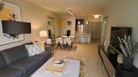 This well-appointed single-floor residence offers a thoughtfully designed open-concept layout that integrates the living, dining, and kitchen areas into one cohesive and inviting space. The living area provides a comfortable gathering space anchored by a contemporary sofa, decorative throw pillows, and a stylish coffee table atop a richly patterned area rug. A large decorative mirror and a curated selection of bold abstract artwork adorn the walls, adding personality and visual depth to the space. The dining area transitions naturally from the living room, featuring a sleek dining table with modern cross-back chairs, ideal for entertaining. Track lighting overhead ensures the space is well-lit and functional. The kitchen is a standout feature, boasting stainless steel appliances, a central island with pendant lighting, and dark cabinetry that contrasts beautifully with the lighter tones of the surrounding space. Neutral-toned carpet flows throughout the main living areas, contributing to a warm and welcoming ambiance. The overall aesthetic is modern and sophisticated, balancing clean lines with carefully chosen decorative elements that create a refined yet livable environment.