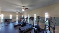 Located at 5500 El Camino del Rey St in Houston, Texas, this fitness center offers a well-equipped, single-floor workout facility designed to support a range of exercise routines. The room features a distinctive two-tone wall treatment, with a warm beige upper half and a cool teal lower half divided by a clean horizontal band, giving the space a calm, structured aesthetic. Dark rubber flooring runs throughout, providing durability and traction underfoot. Multiple arched windows line the walls, flooding the interior with natural light and offering views of the surrounding greenery, creating an open and refreshing atmosphere. Ceiling fans are mounted overhead, ensuring comfortable air circulation during workouts. The cardio section is well-stocked with treadmills, elliptical trainers, and upright stationary bikes from brands including Precor and Cybex. A dedicated free-weight area houses a dumbbell rack alongside a weight tree with medicine balls, complemented by exercise mats for floor-based training. The layout is thoughtfully organized, allowing users to move freely between equipment. An emergency exit door and fire safety equipment are also present, underscoring the facility's attention to safety standards. Overall, the space delivers a functional and well-lit environment suited for daily fitness use.