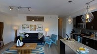 This single-floor space showcases a thoughtfully designed open-concept layout that connects the living and kitchen areas into one cohesive, functional environment. The living area is anchored by sleek hardwood-style flooring and features a ceiling fan, track lighting, and a bold color palette brought to life through contemporary furnishings, including a deep navy sofa, a vibrant teal accent chair, and a graphic blue-and-white area rug. Abstract artwork adorns the crisp white walls, adding a creative touch to the space. A rustic wood coffee table grounds the seating arrangement and provides a pleasing contrast to the modern aesthetic. Natural light filters through the open layout, contributing to the airy and welcoming ambiance. The kitchen area is visible from the living space and is defined by a granite countertop peninsula that serves as both a functional prep space and a casual dining spot. A statement pendant light with a clear glass shade hangs above, adding an elegant focal point. Dark cabinetry in the kitchen creates a bold contrast against the lighter walls and countertops. A hallway leading further into the space hints at additional rooms beyond, completing what is a well-appointed and stylishly curated single-level home.