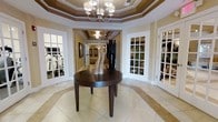 This single-floor amenity space makes a strong first impression with a beautifully appointed foyer at its heart. The entryway features a tray coffered ceiling with warm tonal banding, anchored by a classic multi-arm chandelier that casts a welcoming glow throughout the space. Large format cream and beige tile flooring with inlaid border detailing adds a refined, polished aesthetic underfoot.

A round dark-stained wood accent table sits centrally in the foyer, styled with decorative vases that add an artistic touch to the space. The layout is thoughtfully organized, with multiple French-style glass-paned doors branching off from the main entry. To the left, a well-equipped fitness center is visible, housing cardio equipment behind glass. To the right, a secondary room — likely a lounge or common gathering area — is accessible through matching French doors, offering views of decorative accents and greenery within.

A hallway extends from the foyer deeper into the space, lined with framed artwork and additional tray ceiling details, suggesting further rooms or amenities beyond. The overall ambiance is upscale and professionally designed, combining classical architectural elements with modern comforts to create a welcoming, functional environment suited for community or hospitality use.