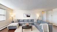 This single-floor residence offers a thoughtfully arranged layout that balances comfort and functionality across its key living spaces. The open-plan living and dining area forms the heart of the unit, where rich hardwood flooring runs throughout and large windows fitted with blinds allow generous natural light to fill the room. The living area accommodates a generous sectional sofa centered around a coffee table, with a large flat-screen TV and ample closet storage nearby. The adjacent dining space features a full-sized rectangular dining table beneath a pendant light fixture, creating a defined yet connected area for meals. The kitchen is compact but well-equipped, showcasing warm cherry wood cabinetry, granite-style countertops, tile flooring, a stainless steel dishwasher, and a gas range — offering everything needed for everyday cooking. Both bedrooms are spacious with hardwood-style flooring, neutral walls, textured ceilings, and room for a full workspace setup. The primary bedroom includes two separate closet doors, providing practical storage. The bathroom presents a clean, all-white aesthetic with subway tile surrounding a full soaker tub and shower combo. Throughout the residence, a consistent neutral color palette and textured ceilings contribute to a calm, livable ambiance.