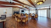 This expansive single-floor gathering space combines warm architectural design with a welcoming, lodge-inspired ambiance. Soaring vaulted ceilings are defined by prominent dark wood beams that extend dramatically across the full length of the room, complemented by a white-paneled ceiling and ceiling fans that enhance air circulation throughout. Rich hardwood flooring runs seamlessly across the open layout, grounding the space in warmth and elegance.

A striking stone fireplace serves as a natural focal point on one end of the main room, framed by a flat-screen television above the mantle, making it both a functional and aesthetic centerpiece. Multiple seating arrangements — featuring upholstered armchairs, side tables, and a glass-top coffee table — are thoughtfully arranged to encourage conversation and relaxation, while dining tables with cane-back chairs accommodate group gatherings.

Floor-to-ceiling windows dressed in sheer and patterned drapes line the perimeter, flooding the interior with soft, diffused natural light and offering views of the surrounding greenery. Elegant chandeliers and table lamps add layered lighting throughout. A tiled entryway foyer, visible through an arched passageway, provides a gracious entry point into the space, complete with its own chandelier and access to exterior doors.