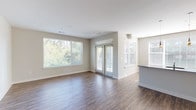 The Hawkins unit offers a well-designed single-story layout accommodating three bedrooms and three full bathrooms. The open living area immediately stands out, featuring rich hardwood-style flooring and an abundance of natural light streaming through multiple large windows fitted with blinds. French doors lead to an outdoor view of mature trees, enhancing the bright, airy ambiance of the space. A peninsula counter separates the living area from the kitchen, which is outfitted with light maple cabinetry, dark gray countertops, and a full suite of black appliances including a refrigerator, range, and built-in microwave. Track lighting and a large window complete the kitchen's functional design. The bedrooms feature plush neutral carpeting and generous windows that invite natural light and outdoor views. Each bedroom benefits from a walk-in closet equipped with wire shelving systems, providing ample storage. The bathrooms are finished consistently throughout, showcasing light maple vanities topped with dark countertops, undermount sinks, wood-look flooring, and tiled tub-shower combinations. An in-unit laundry area is also present, adding everyday convenience to this well-appointed residence.