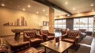 Located at 548 Newtown Rd in Virginia Beach, VA, this single-floor amenity space blends modern industrial design with warm, rustic elements to create an inviting and multifunctional environment. Light wood-plank flooring flows throughout, unifying the distinct zones of the space.

The lounge area is a standout feature, anchored by a striking reclaimed wood accent wall and outfitted with tufted leather armchairs arranged around low tables. Two wall-mounted flat-screen TVs add functionality, while large windows fitted with roller shades and patterned curtains allow natural light while maintaining privacy. Exposed black ductwork runs across the ceiling, lending an industrial character that complements the warm furnishings.

Adjacent to the lounge is a game room area with a shuffleboard table, bar-height seating, and open floor space — ideal for leisure and social gatherings.

The café-style dining section features long communal tables with modern saddle-brown chairs lined along a wainscoted wall adorned with eclectic decorative arrangements, including framed art, greenery, and wall clocks. French doors offer views and access to an outdoor area, filling the room with natural light.

A central bar and service counter runs the length of the space, featuring butcher block countertops, dark cabinetry, a sink, beverage refrigerators, and rustic wood accents — providing a fully functional hosting hub for residents and guests alike.