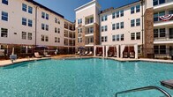 Located at 3171 US-280 E in Birmingham, Alabama, this outdoor amenity space is designed to deliver a resort-like experience within a modern residential community. The centerpiece is a generously sized swimming pool with crystal-clear blue water, complemented by a separate spa area that encourages relaxation. The pool deck is furnished with lounge chairs positioned to maximize sun exposure, while elegant white-curtained private cabanas line one side of the deck, offering shaded retreats for residents. Wooden pergolas anchor additional seating areas, providing a covered outdoor dining and lounging environment framed by mature trees and manicured landscaping. A distinctive natural flagstone patio area adds a rustic yet refined touch, enclosed by sleek black iron fencing that defines the space without obscuring the surrounding greenery and architectural views. The multi-story building surrounding the courtyard features a classic exterior combining cream-colored siding with warm stone accent towers and private balconies that overlook the amenity area below. The overall ambiance is open and sun-drenched, with thoughtful landscaping softening the built environment and creating a welcoming, community-centered outdoor setting ideal for both active recreation and leisurely enjoyment.