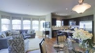 This single-floor residence showcases a thoughtfully designed open-concept layout that effortlessly connects the living, dining, and kitchen areas into one cohesive and functional space. Rich hardwood flooring flows throughout, anchoring the warm yet contemporary aesthetic.

The living area is bathed in natural light streaming through a series of arched windows dressed with decorative valances, creating an inviting and airy atmosphere. The space comfortably accommodates a seating arrangement centered around a soft area rug, offering a relaxed yet refined gathering spot.

The dining area features a sleek glass-top table with upholstered seating, positioned to take full advantage of the open sightlines across the space. Adjacent to the dining area, the kitchen is appointed with dark espresso cabinetry, light countertops, and stainless steel appliances, including a microwave, dishwasher, and range. A breakfast bar with seating provides additional casual dining options and further integrates the kitchen into the social flow of the home.

Recessed lighting supplements the abundant natural light, ensuring the space remains bright and welcoming at all times. The neutral color palette of soft grays and warm taupes throughout lends a clean, modern sensibility, while the overall layout maximizes both comfort and practicality.