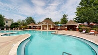 Located at 2000 Geddy Ln in Cary, North Carolina, this single-story community amenity facility serves as a central outdoor gathering hub defined by its resort-style atmosphere and thoughtfully designed grounds. The centerpiece is a generously sized, freeform swimming pool with clear turquoise water, complemented by a secondary pool area ideal for relaxing. Surrounding the pools, an expansive brick-paver pool deck provides ample space for lounge chairs and umbrellas, creating a comfortable environment for outdoor recreation.

The clubhouse structure features classic Southern architectural details, including a covered porch with tall white columns, ceiling fans, and rocking chairs that invite leisurely relaxation. The building's exterior combines cream-colored siding with brick accents and a pitched, shingled roof with distinctive pavilion-style covered areas offering shaded seating and dining options near the pool.

Mature trees, manicured shrubs, colorful flower beds, and decorative stone landscaping elements frame the entire area, enhancing the natural ambiance. A secured, wrought-iron gate with a keypad entry system ensures controlled access to the facility. The overall setting presents a polished, well-maintained outdoor environment suited for community gatherings and leisure activities throughout the warmer months.