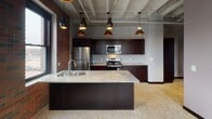 Located at 204 E Grand Ave in downtown Eau Claire, Wisconsin, this single-floor unit combines industrial character with modern finishes to create a distinctive urban living experience. The open-concept kitchen serves as the centerpiece of the space, showcasing rich dark cabinetry, granite countertops, and a substantial center island with a built-in sink — ideal for both meal preparation and casual entertaining. Stainless steel appliances, including a refrigerator, electric range, and over-the-range microwave, complete the well-appointed kitchen. Dramatic pendant lighting with copper-toned interiors hangs above the island, adding warmth to the space. Exposed brick walls run along one side of the kitchen, lending an authentic industrial aesthetic that is complemented by the polished terrazzo flooring found throughout. The soaring ceilings feature exposed structural beams and visible pipe work, a hallmark of loft-style architecture. Natural light filters in through large black-framed windows, offering views of the surrounding downtown streetscape. Additional rooms include a clean, functional bathroom with a full tub/shower combination, and a versatile secondary room with neutral walls and recessed lighting — well-suited as a bedroom or flex space. The overall ambiance is bold yet refined, blending raw architectural elements with contemporary upgrades.