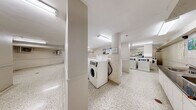 Located at 25 Saint Mary Street in the heart of Toronto, this shared laundry facility offers a well-organized and functional space for residents. The floor plan is thoughtfully laid out across a single level, providing a generous and open area designed to accommodate multiple users at once.

The laundry room is the centerpiece of the space, equipped with a row of modern coin-operated washing machines and dryers arranged in a clean, efficient configuration. The machines are clearly labeled and accompanied by posted instructions, ensuring ease of use. Bright fluorescent lighting illuminates the room evenly, creating a well-lit environment throughout.

The flooring throughout the entire facility consists of classic speckled terrazzo, which is both durable and easy to maintain. The walls are finished in a neutral off-white palette, contributing to a clean and utilitarian aesthetic.

Adjacent to the laundry area is a comfortable waiting room, furnished with a glass-top table and chairs, providing residents with a place to relax while their laundry cycles complete. A corridor connects the various sections of the facility, lined with informational notices and leading to the main entrance door.

Climate control units are mounted on the walls to ensure year-round comfort. A payment kiosk near the entrance adds a convenient, centralized method for managing laundry transactions. Overall, this facility is practical, tidy, and well-equipped to serve a residential building community.