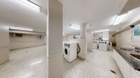 Located at 25 Saint Mary Street in the heart of Toronto, this shared laundry facility offers a well-organized and functional space for residents. The floor plan is thoughtfully laid out across a single level, providing a generous and open area designed to accommodate multiple users at once.

The laundry room is the centerpiece of the space, equipped with a row of modern coin-operated washing machines and dryers arranged in a clean, efficient configuration. The machines are clearly labeled and accompanied by posted instructions, ensuring ease of use. Bright fluorescent lighting illuminates the room evenly, creating a well-lit environment throughout.

The flooring throughout the entire facility consists of classic speckled terrazzo, which is both durable and easy to maintain. The walls are finished in a neutral off-white palette, contributing to a clean and utilitarian aesthetic.

Adjacent to the laundry area is a comfortable waiting room, furnished with a glass-top table and chairs, providing residents with a place to relax while their laundry cycles complete. A corridor connects the various sections of the facility, lined with informational notices and leading to the main entrance door.

Climate control units are mounted on the walls to ensure year-round comfort. A payment kiosk near the entrance adds a convenient, centralized method for managing laundry transactions. Overall, this facility is practical, tidy, and well-equipped to serve a residential building community.