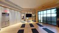 This single-floor fitness studio is thoughtfully designed to support a variety of exercise and wellness activities. The main exercise area features light hardwood flooring, providing a clean and warm foundation for the multiple yoga mats and step platforms arranged throughout the space. Recessed lighting is embedded in a subtly tray ceiling, casting an even, welcoming glow across the room.

A wall-mounted flat-screen television is centrally positioned within a built-in niche flanked by large mirrors, allowing participants to monitor their form during workouts. The mirrors also enhance the sense of depth and openness in the room. Adjacent to the TV niche, an open doorway provides a view into a secondary space, suggesting additional amenities or rooms beyond.

A floor-to-ceiling window wall with a dark metal frame lines one side of the studio, introducing abundant natural light and offering views of the surrounding outdoor environment. Soft, neutral wall tones and warm accent lighting contribute to a calm, focused ambiance well-suited for yoga, stretching, or group fitness classes. The overall design balances functionality with a serene aesthetic.