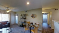 This single-floor residence offers a thoughtfully arranged open-concept layout that connects key living areas with ease and flow. The dining area serves as a focal point of the space, featuring a round glass-top table surrounded by contemporary tan chairs, complemented by a decorative wrought-iron baker's rack that adds a classic, ornate touch. A circular wall art piece adds a modern accent against the warm, neutral-toned walls. Recessed lighting overhead ensures the space is well-lit, while a door with a sidelight panel invites generous natural light into the area, enhancing the bright and airy atmosphere. The flooring transitions seamlessly from plush carpeting in the dining and living zones to rich hardwood flooring visible in the adjacent room, providing both comfort and visual contrast. A peek into the connecting room reveals additional natural light streaming through large windows, suggesting an equally welcoming space beyond. The overall ambiance is warm and inviting, with a neutral color palette that creates a versatile backdrop suitable for a variety of interior styles. The layout is efficient and well-proportioned, making excellent use of the available space across the single floor.