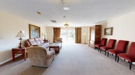 Located at 414 Berckmans Rd in Augusta, Georgia, this single-floor commercial space offers a well-organized layout suited for professional gatherings or community-oriented use. The main gathering area is generously proportioned, featuring plush wall-to-wall carpeting, a textured ceiling, and warm neutral walls that create a calm, welcoming atmosphere. Ornate flush-mount light fixtures provide ambient illumination throughout, while large French doors and windows dressed in rich gold drapes allow considerable natural light and offer views of the surrounding outdoor grounds.

The open layout of the main area accommodates multiple seating arrangements, with distinct zones that include a comfortable lounge area and a more formal arrangement with upholstered seating and wooden storage furnishings. A private office area is equipped with a traditional executive desk and side chairs, framed artwork adding a refined touch.

A galley-style kitchen features light wood cabinetry, white countertops, tile flooring, and a full suite of black appliances including a refrigerator, range, microwave, and dishwasher. An adjacent utility corridor with additional cabinetry and countertop workspace connects back to the main area. The space also includes an accessible restroom with tile flooring and grab bar installations, ensuring compliance and convenience for all users.