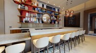 This bar lounge is a thoughtfully designed social space that seamlessly blends upscale aesthetics with a welcoming atmosphere. The centerpiece is a full-service bar topped with a sleek white surface, framed by warm wood shelving that spans the wall behind it. The shelving displays decorative ceramic vessels set against a vibrant, geometric mural in bold reds, blues, and yellows, creating a striking visual backdrop. Copper globe pendant lights hang overhead, adding a warm, ambient glow. White upholstered bar stools with gold-toned legs line the bar counter, offering comfortable seating.

Beyond the bar, the lounge opens into generously proportioned seating areas. Plush yellow accent chairs, a striped sofa, and a low glass coffee table are arranged to encourage conversation, while a round glass-top bistro table provides an intimate nook for smaller gatherings. Large-scale abstract artwork punctuates the neutral walls throughout.

A distinctly appointed alcove, framed by rich wood paneling and lit by a chandelier, features a padded banquette and ottomans for a more private lounge experience. Floor-to-ceiling windows and glass entry doors flood the space with natural light and provide views of an outdoor courtyard, creating an effortless connection between the interior and exterior.