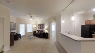 This single-floor residence offers an open and airy layout designed for comfortable everyday living. The main living area is finished with soft, wall-to-wall carpeting and features a ceiling fan that contributes to a relaxed atmosphere. Natural light filters in through blinds-covered windows, brightening the neutral, warm-toned walls throughout.

Adjacent to the living room, the kitchen is defined by a sleek countertop peninsula that serves as both a functional workspace and a casual dining or entertaining surface. Stylish pendant lights hang above the peninsula, adding a modern touch to the space. The kitchen is equipped with contemporary fixtures including a clean, modern faucet and ample counter space.

An arched doorway leads from the living area into what appears to be a hallway connecting to additional rooms, providing a sense of depth and flow throughout the unit. The open sightlines between the kitchen and living areas create a seamless, connected feel that is both practical and inviting.

Overall, the space presents a clean, well-maintained interior with a neutral color palette, modern lighting accents, and a functional layout that balances openness with defined living zones.