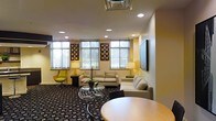 This single-floor space presents a well-appointed lounge area designed with both comfort and functionality in mind. The room is illuminated by a combination of recessed ceiling lights and warm table lamps, creating a balanced and inviting ambiance throughout. Large windows along one wall allow generous amounts of natural light to filter in, offering views of the surrounding greenery outside.

The seating area is furnished with contemporary neutral-toned sofas and a distinctive accent chair in a bold green hue, adding a pop of color to the otherwise understated palette. A sleek glass-and-chrome coffee table serves as a central focal point, complementing the modern aesthetic of the space. Decorative wall art featuring geometric square patterns is displayed between the windows, adding visual interest to the cream-colored walls.

A large flat-screen display is mounted on the right-side wall, suggesting the space supports presentations or entertainment. A round wood-toned table and chairs are partially visible in the foreground, indicating a dedicated area for seated gatherings or collaborative work. The patterned carpet anchors the room with a dark, graphic design that contrasts with the lighter furnishings, contributing to the space's polished, professional character.