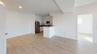 This single-floor unit offers a clean, contemporary design characterized by crisp white walls, light wood-toned flooring, and recessed lighting throughout. The open-concept living and kitchen area creates a seamless, airy flow, anchored by a peninsula that defines the kitchen space without sacrificing openness. The kitchen is well-appointed with white shaker-style cabinetry, dark contrasting countertops, stainless steel appliances including a gas range, over-the-range microwave, and refrigerator, all complemented by brushed nickel hardware. The living area is generously proportioned and benefits from abundant natural light, with views of the surrounding neighborhood visible through large windows. Multiple bedrooms feature the same light hardwood flooring, bright white walls, and windows that invite natural light. The bathroom is thoughtfully finished with a wide-format vanity mirror, white quartz countertop, grey cabinetry, and a combination shower-tub enclosed by sleek sliding glass doors. A dedicated in-unit stacked washer and dryer adds everyday convenience. The overall ambiance is modern, bright, and move-in ready, with a neutral palette that offers versatile styling potential.